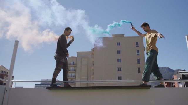 Two men navigate a rooftop edge holding bright flares against a cityscape backdrop. The vibrant colors of the flares contrast with the blue sky, capturing a moment of urban adventure. Suitable for themes of adrenaline-fueled sports, urban exploration, and youthful creativity.