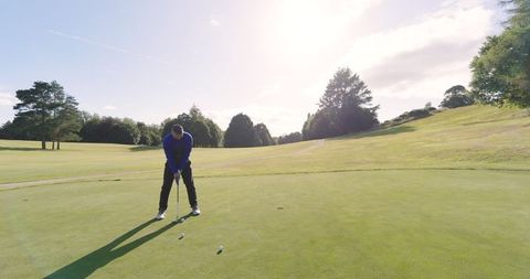 A mid adult male golfer is seen bending over at a putting green, intently gripping his putter and lining up a golf ball close to the hole. The vast, well-maintained golf course stretches towards the sky, enveloped in bright sunlight. This scene encapsulates relaxation and mindfulness in sport. Ideal for content centered on golf, sports commercials, outdoor tourism, or promotional materials for golfing tournaments.
