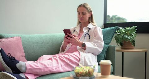 Female medical professional relaxing on clinic sofa in pink scrubs checking smartphone