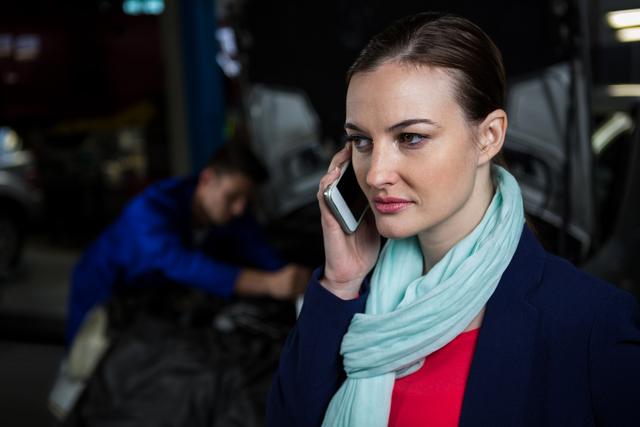 Businesswoman standing in repair garage speaking on mobile phone while auto mechanic works in the background. Ideal for use in automotive services, customer service, communication, and business-related projects.