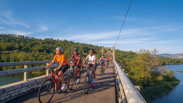 Cyclists riding across scenic river bridge, road riders in colorful jerseys and helmets