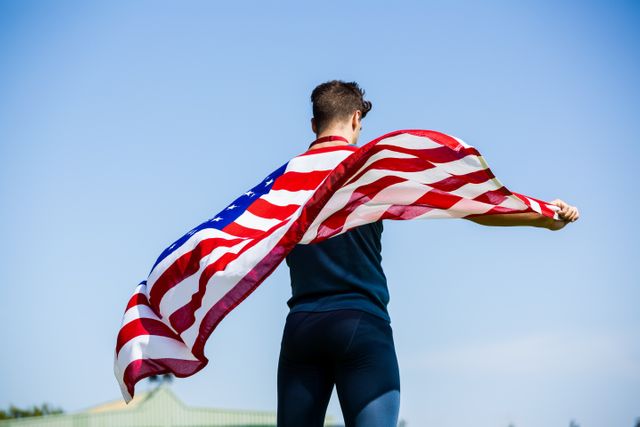 This image shows an athlete holding an American flag outdoors, symbolizing patriotism and national pride. The clear blue sky adds to the sense of freedom and celebration. Ideal for use in campaigns related to sports, national holidays, fitness, and unity.