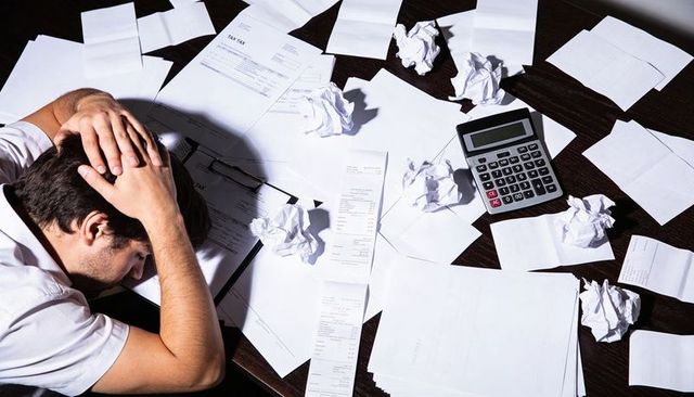Overwhelmed man holding head at cluttered desk with bills, calculator, crumpled receipts