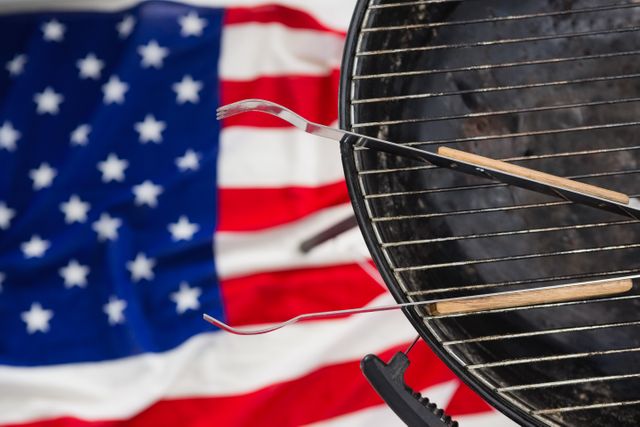 Barbecue grill with tongs placed on it, with an American flag in the background. Ideal for use in content related to summer cookouts, patriotic holidays like Independence Day, and outdoor cooking events. Perfect for illustrating themes of American culture, celebrations, and family gatherings.