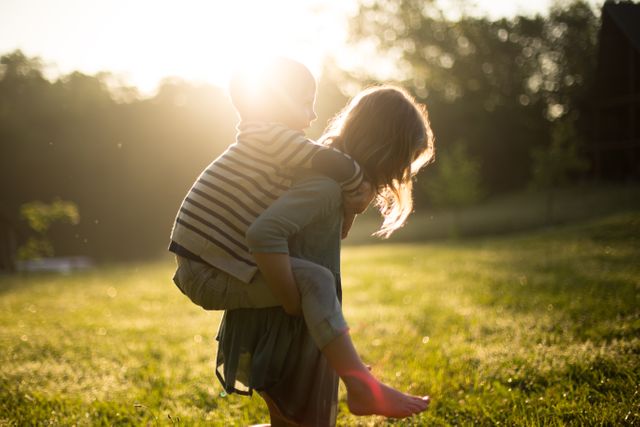 Siblings enjoying fun time together in a sunlit park with a piggyback ride. Useful for themes about family bonding, outdoor activities, childhood memories, and happiness. Perfect for advertisements, blog posts, or social media posts targeting family-oriented audiences.