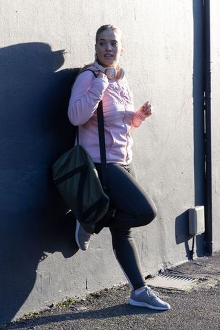 Young woman in pink hoodie leaning on urban wall with duffel bag and headphones