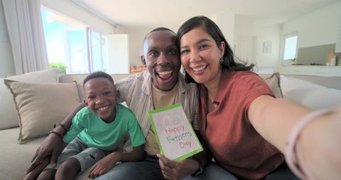 Smiling family taking selfie on sofa holding handmade happy fathers day card