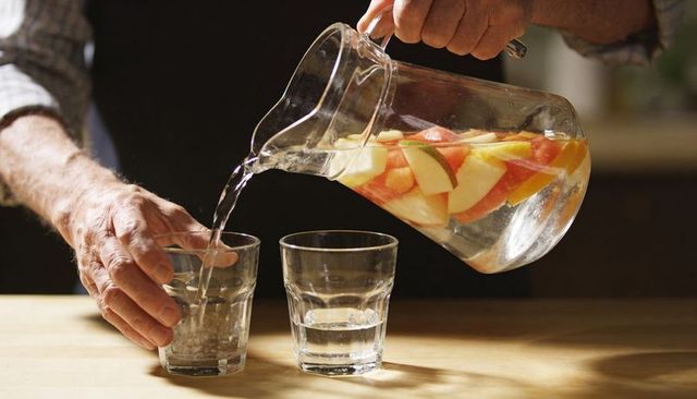 Senior hands pouring fruit-infused water from glass pitcher into tumblers on wooden table