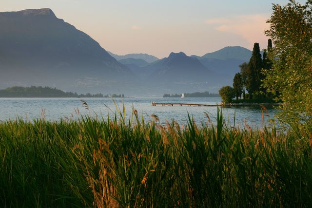 Tranquil scene featuring a lake with a distant dock, surrounded by lush grass and mountains in the background at sunset. Ideal for travel guides, environmental content, landscape photography collections, or promotional material for nature retreats.