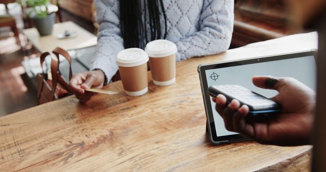 Image showing two customers at a cozy coffee shop counter, making a purchase. One is using a credit card, while the other uses a smartphone for payment. This image can be used for illustrating concepts like digital payments, customer convenience, or modern technology in retail industry.