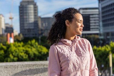 Young woman squinting in morning sun on urban rooftop wearing pink quilted hooded jacket