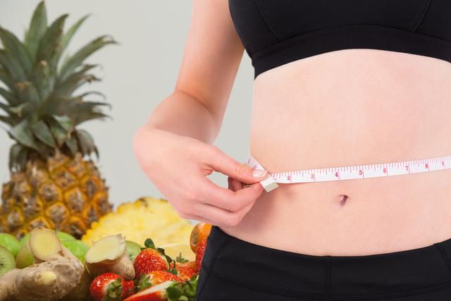 Midsection of woman measuring waist, surrounded by variety of fresh fruits including pineapple, ginger, and strawberries. Ideal for use in articles about health, fitness, diet plans, healthy eating, fresh produce benefits, and exercise routines.