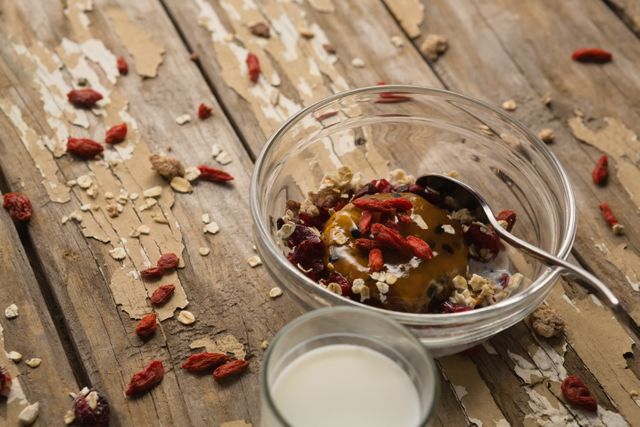 This image shows a healthy breakfast setup with cereal, goji berries, and oats in a glass bowl, accompanied by a glass of milk on a rustic wooden table. Ideal for use in articles or advertisements related to healthy eating, breakfast recipes, nutrition, and diet plans. It can also be used in blogs or social media posts promoting wholesome morning meals and superfoods.