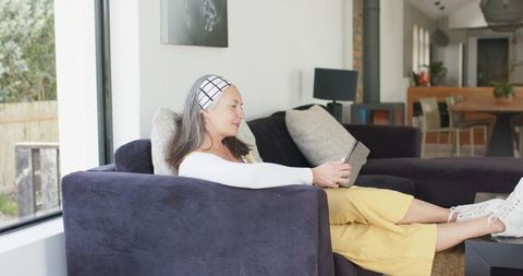 Mature woman relaxing on dark sofa holding tablet in sunlit modern living room