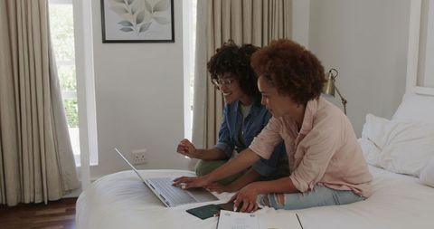 Friends planning trip on laptop while sitting on bed with passport and travel documents