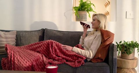 Young woman relaxing on sofa, speaking into smartphone, wrapped in red plush blanket