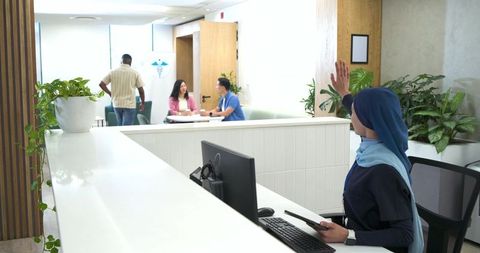 Clinic receptionist in navy hijab welcoming visitors while holding tablet at modern desk