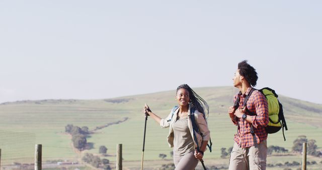 Young couple hiking together in a scenic rural landscape. The pairs are carrying backpacks and using walking sticks. Ideal for promoting outdoor activities, travel adventures, and healthy lifestyles. Use for advertising hiking gear, travel destinations, or fitness-related content.