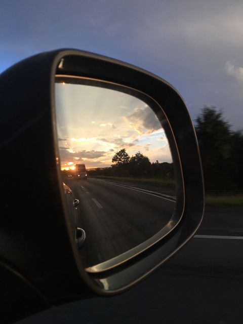 A vivid sunset reflects in the car side mirror while driving on a highway. The image captures the golden hour as it blends with road travel. Ideal for themes related to travel, adventure, road trips, the beauty of nature in everyday moments, reflective thinking, and memories. It is perfect for blogs, articles, or campaigns focusing on journeying, capturing precious moments on the go, or evoking a sense of exploration and freedom.