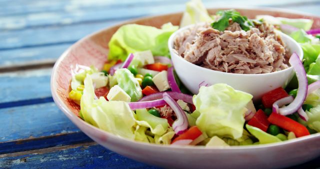 This vibrant image showcases a healthy tuna salad brimming with fresh vegetables such as lettuce, red bell peppers, peas, corn, and onions, served in a bowl on a rustic wooden table. Perfect for promoting balanced diets, meal planning, culinary blogs, and healthy eating campaigns.