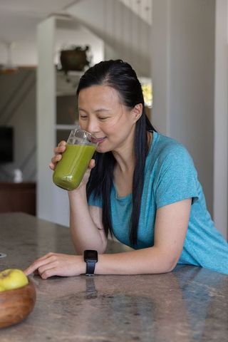 Asian woman sipping green smoothie while leaning on kitchen counter, wellness lifestyle