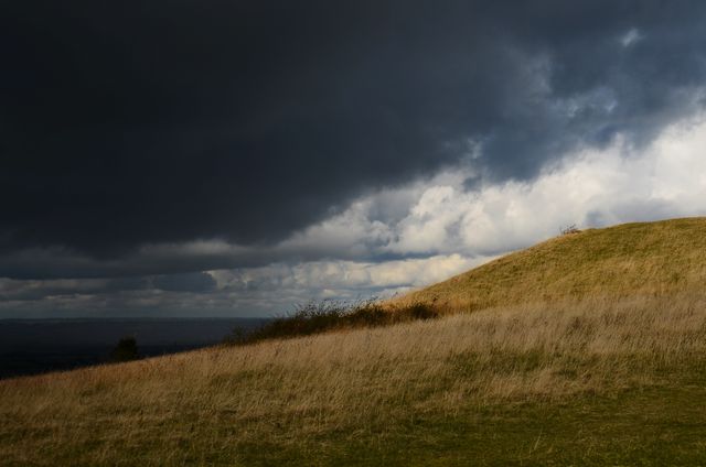 Stormy sky covering rolling hills with dry grass on a cloudy day, creating a dramatic contrast between light and dark. This scene can illustrate weather changes, natural outdoors, and landscape photography themes. Ideal for nature calendars, weather-related articles, and geographical studies.