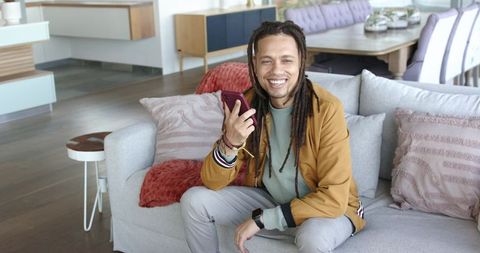 Smiling man with dreadlocks using smartphone on modern living room sofa, relaxed lifestyle