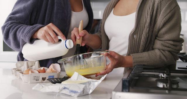 Two women are mixing cake batter in a modern kitchen, with milk and eggs on the counter. This can be used for content related to cooking, baking, home life, family activities, and togetherness.
