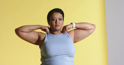 Confident midlife woman standing wearing blue tank top and smartwatch on yellow-gray backdrop