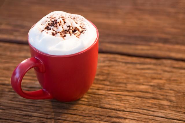 Red mug filled with coffee topped with whipped cream and chocolate sprinkles on wooden table. Perfect for use in holiday promotions, coffee shop advertisements, and cozy winter-themed designs.