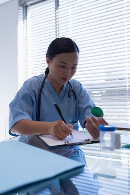 Female doctor wearing a stethoscope around her neck, sitting at a desk in a clinic, writing a prescription on a clipboard. She is focused on her task, holding a medicine bottle. Useful for depicting healthcare professionals, medical procedures, hospital settings, and clinical environments.