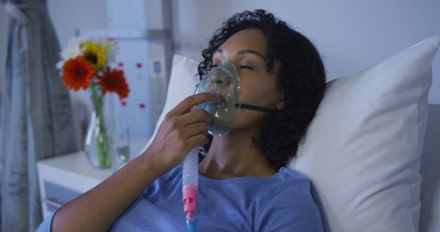 This image features a woman in a hospital gown lying in a bed, using an oxygen mask. The setting includes a floral arrangement on a bedside table. Useful for healthcare, medical, recovery, treatment-related content. It can be used in articles, healthcare advertising, or educational materials illustrating patient care and recovery.
