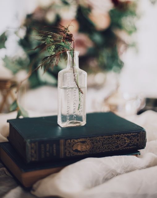 This cozy image features a vintage-style arrangement with an old glass bottle containing a plant, delicately placed on top of antique books. The blurred floral background and white fabric add a soft, inviting atmosphere. Perfect for use in blogs, websites, or media projects related to home decor, vintage collections, and aesthetic inspiration.