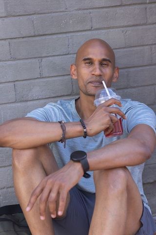 African american man sitting and sipping iced drink against urban gray brick wall
