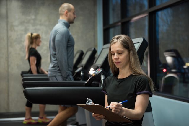 Woman writing on clipboard with people exercising on treadmill at gym