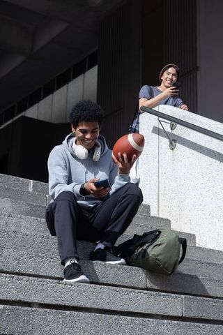 African american and asian teens sitting on campus steps holding football and smartphones