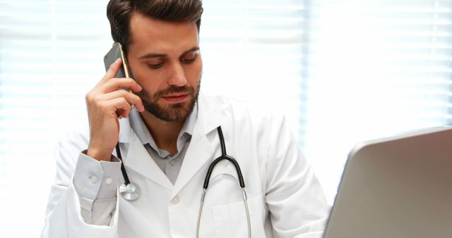 A young Caucasian male doctor is engaged in a conversation on his mobile phone while looking at his laptop, with copy space. His focused expression and stethoscope suggest he is discussing a patient's case or coordinating care.