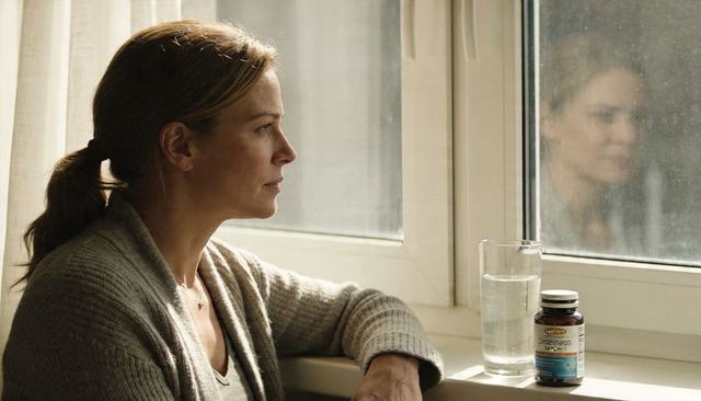 Woman gazing out window in cardigan with supplement bottle and glass, contemplative moment