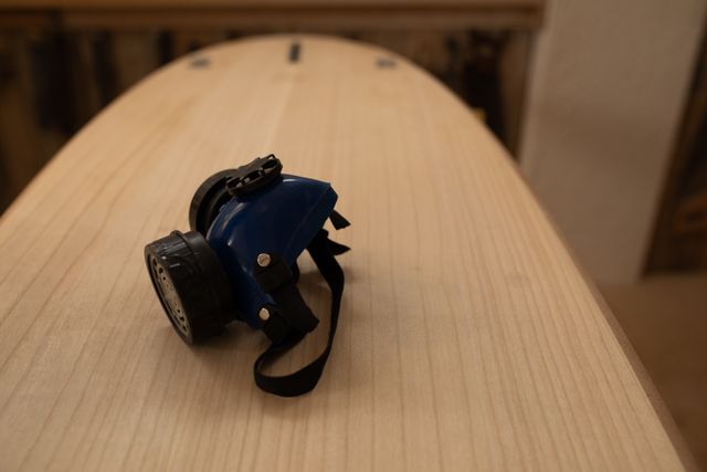 The safety breathing mask of a surfboard maker lying on the smooth wooden surface of a surboard being made on a workbench in a workshop, after shaping with a sander.