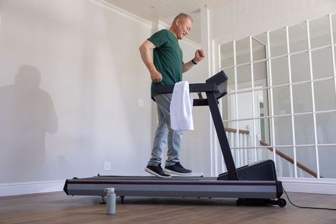 Mature chinese man briskly walking on treadmill at home gym with towel and water bottle