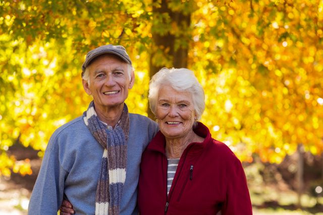 Elderly couple smiling and standing close together in a park during autumn. Ideal for use in advertisements, brochures, or articles related to senior living, retirement, health and wellness, and outdoor activities for seniors.
