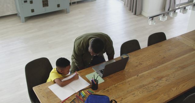 Father assisting son with homework at a large wooden table in a modern living room. Both are focused on the task, with various school supplies on the table. Ideal for educational content, family support, learning concepts, and home environment themes.