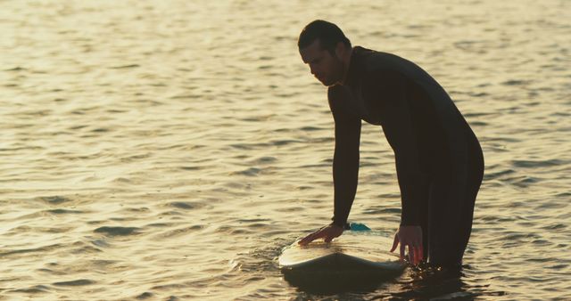 A surfer preparing for a session, standing in the shallow water in evening sunlight. The image captures the serene mood and dedication to the sport. Ideal for use in advertisements for surfing gear, motivational posters, adventure travel blogs, or active lifestyle magazines.