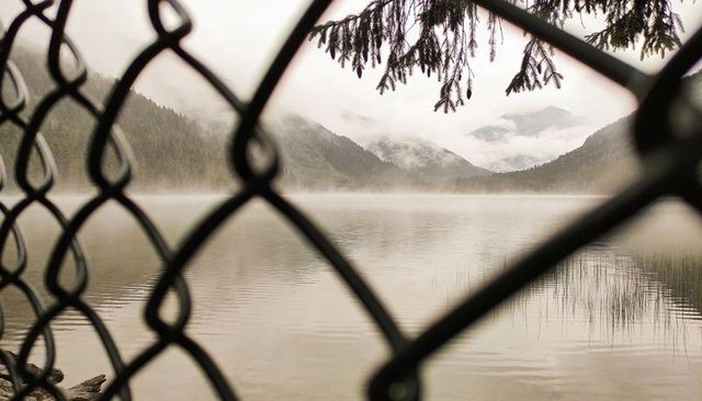 Chain-link fence framing misty alpine lake with calm water, reed beds, forested mountain valley