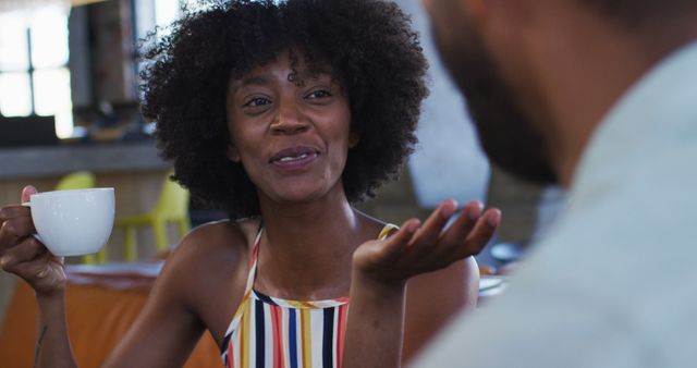 African American woman smiling and engaging in friendly conversation while holding a white coffee cup across a cafe table. Ideal for depicting social interactions, casual meetings, coffee shop atmosphere, relaxation, and friendships.