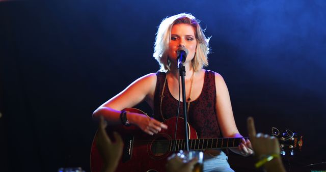 An image of a woman playing a red acoustic guitar and singing into a microphone on stage. The setting is a concert hall with a focused beam of light on the performer. The audience's hands are visible in the foreground, adding to the live music atmosphere. This image can be used for promoting live music events, articles on musicians, or in advertisements for music venues and concerts.