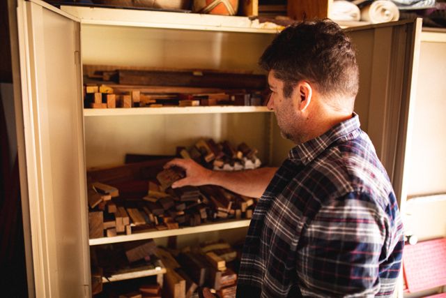Caucasian male knife maker searching for tools in his workshop. He is wearing a plaid shirt and is surrounded by shelves filled with various wood pieces. This image can be used to depict small business, craftsmanship, and manual work. Ideal for articles or advertisements related to woodworking, knife making, DIY projects, and independent artisans.