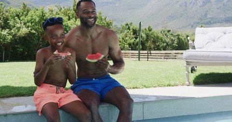 African father and son laughing and eating watermelon while sitting on pool edge in sunny backyard