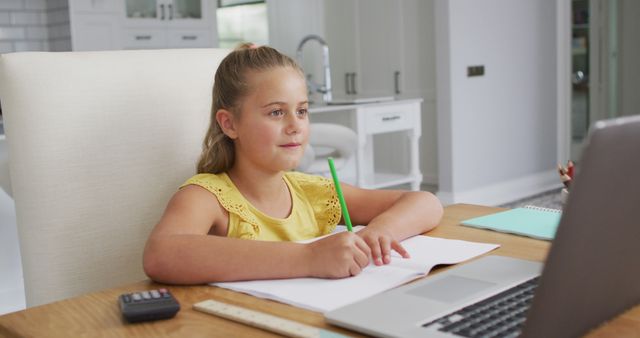 Young girl is sitting at a desk in a modern home setting while studying on a laptop. She is taking notes in a notebook, concentrating on the screen. Scene emphasizes online education, learning, and homework. Useful for topics related to virtual classes, homeschooling, education technology, and childhood learning activities.