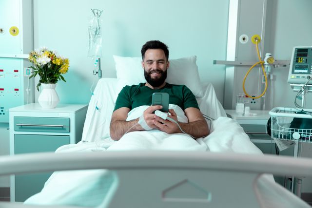 Smiling, bearded caucasian male patient sitting up in hospital bed using smartphone. Medical services, hospital and healthcare concept.
